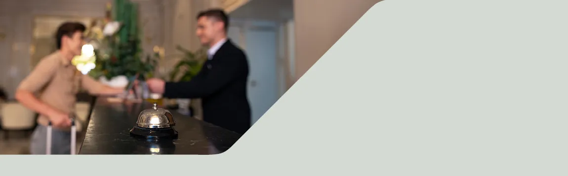 Hotelrepzeption mit einem Gast und einem Hotelangestellten im Hintergrund, im Vordergrund steht eine silberne Empfangsglocke auf den Tresen