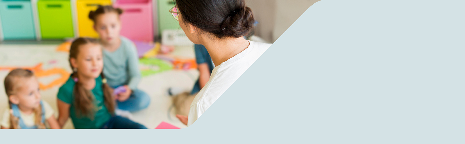 Im Hintergrund sitzen einige Kleinkinder auf dem Boden, im Vordergrund sitzt eine Frau, die zu ihnen spricht