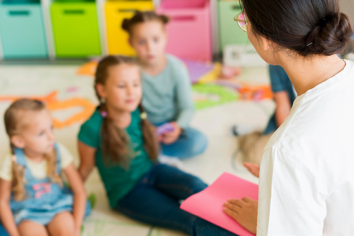 Im Hintergrund sitzen einige Kleinkinder auf dem Boden, im Vordergrund sitzt eine Frau, die zu ihnen spricht