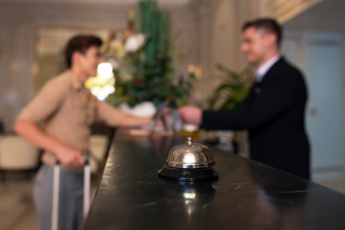 Hotelrepzeption mit einem Gast und einem Hotelangestellten im Hintergrund, im Vordergrund steht eine silberne Empfangsglocke auf den Tresen