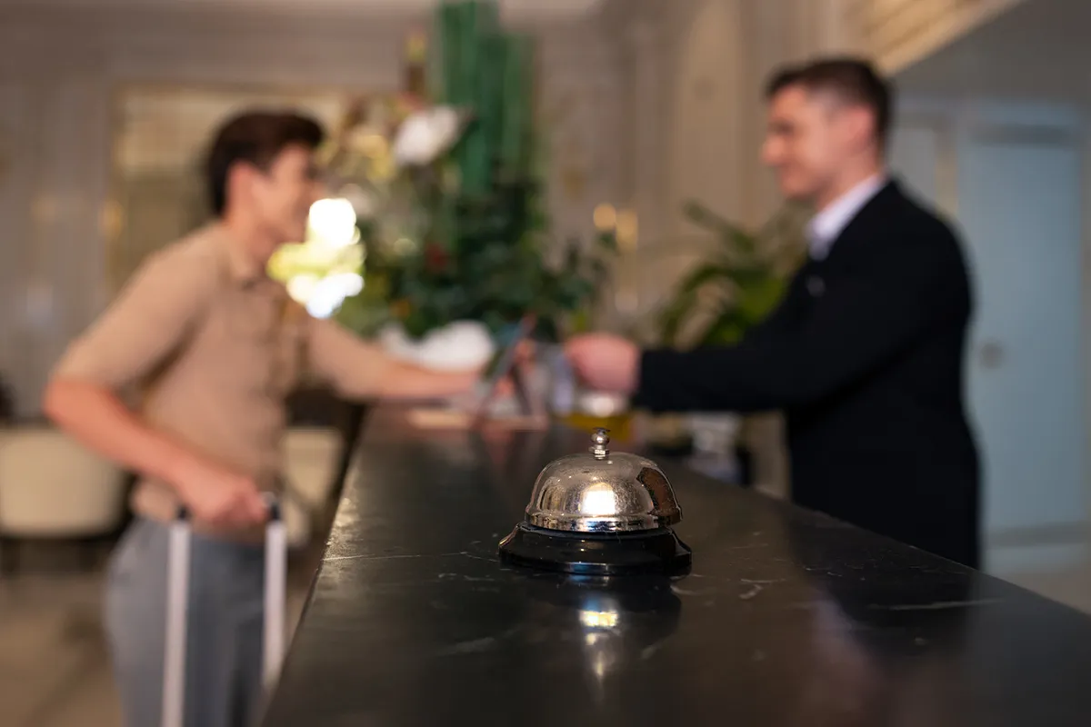 Hotelrepzeption mit einem Gast und einem Hotelangestellten im Hintergrund, im Vordergrund steht eine silberne Empfangsglocke auf den Tresen