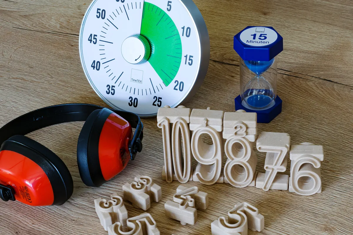 Auf einen Tisch liegen ein roter Gehörschutz eine Zeitdauer-Uhr und eine Sanduhr